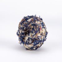 A spherical herbal tea or herbal supplement ball made of dried blue flowers, white wax, and brown herbal ingredients, centered on a clean white background.