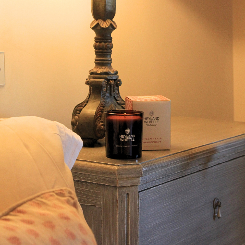 A Heyland Butterfly scented candle in a dark amber glass jar sits on a gray wooden nightstand next to its pink and white product box, with a bed and a large decorative lamp base in the background.