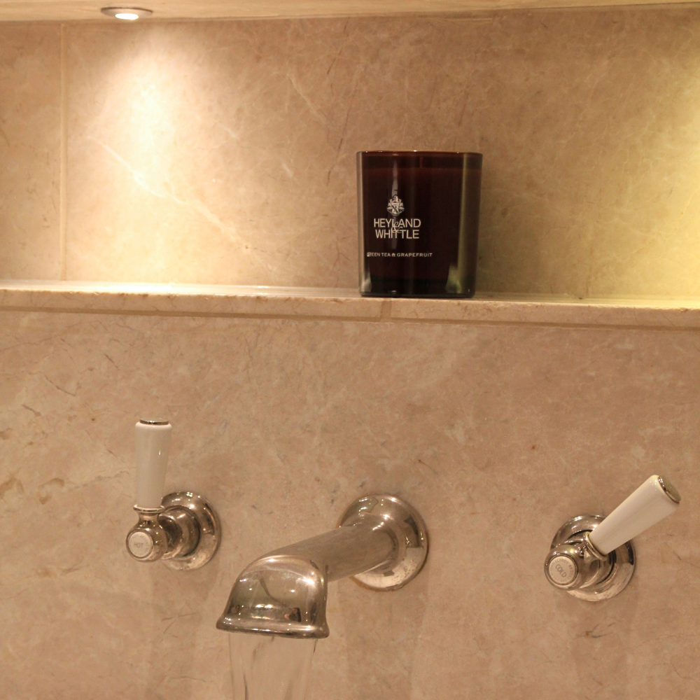 A dark amber glass candle with white text reading 'HEYLAND WATTLE' sits on a beige marble shelf above a chrome faucet in a warm-toned bathroom setting.