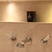 A dark amber glass candle with white text reading 'HEYLAND WATTLE' sits on a beige marble shelf above a chrome faucet in a warm-toned bathroom setting.