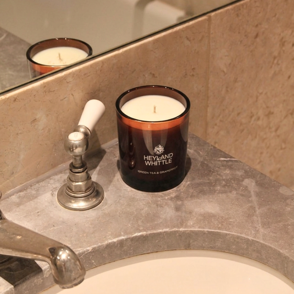 A scented candle in an amber glass jar sits on a bathroom sink counter beside a chrome faucet. The candle has white wax and visible branding that reads 'HEYANO WHITTLE' with 'GREEN TEA & GOURDOPURIS' below.