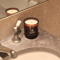 A scented candle in an amber glass jar sits on a bathroom sink counter beside a chrome faucet. The candle has white wax and visible branding that reads 'HEYANO WHITTLE' with 'GREEN TEA & GOURDOPURIS' below.