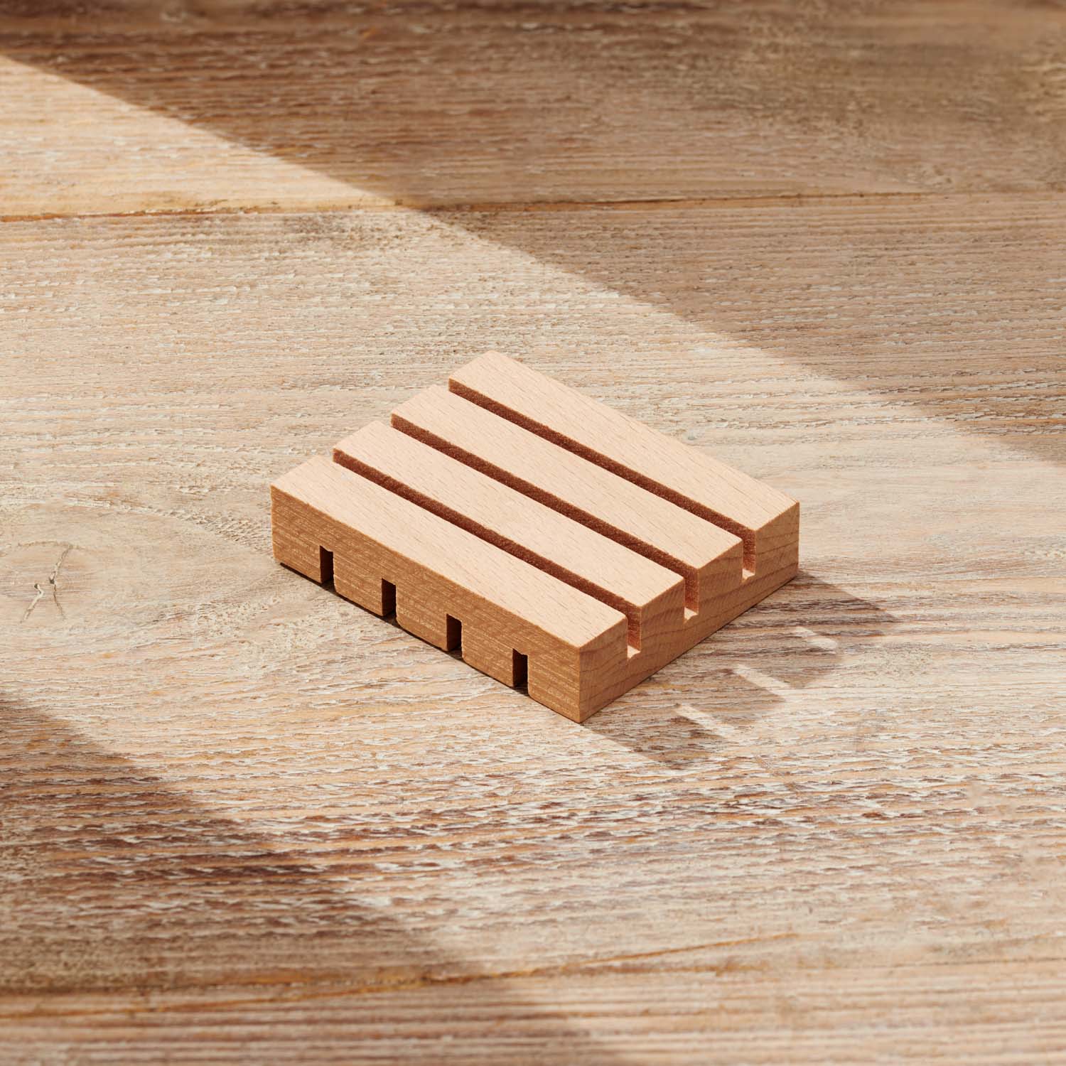 A small rectangular wooden block with four parallel grooves and four rectangular cutouts on one side, photographed on a wood-grain surface with soft diagonal shadows.