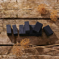 A set of dark charcoal rectangular soap bars arranged on a rustic wooden surface, styled with dried dandelion flowers.