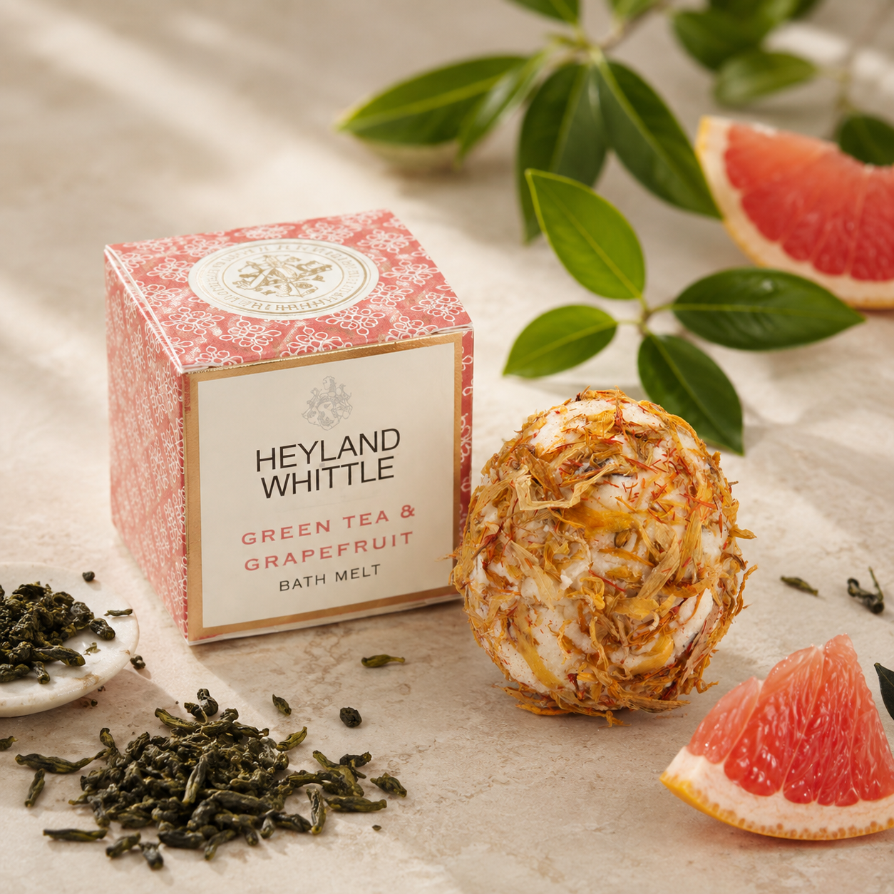 Heyland Whittle Green Tea & Grapefruit Bath Melt in a pink patterned box, styled with a grapefruit ball, loose green tea leaves, and grapefruit slices on a textured beige fabric background.