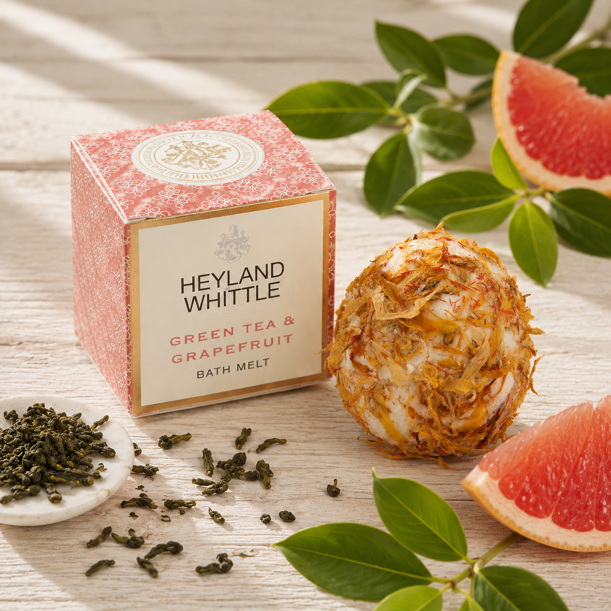 Heyland Whittle Green Tea & Grapefruit Bath Melt shown with its pink patterned box, a bath melt ball, loose tea leaves, grapefruit slices, and green tea leaves on a light wood surface.