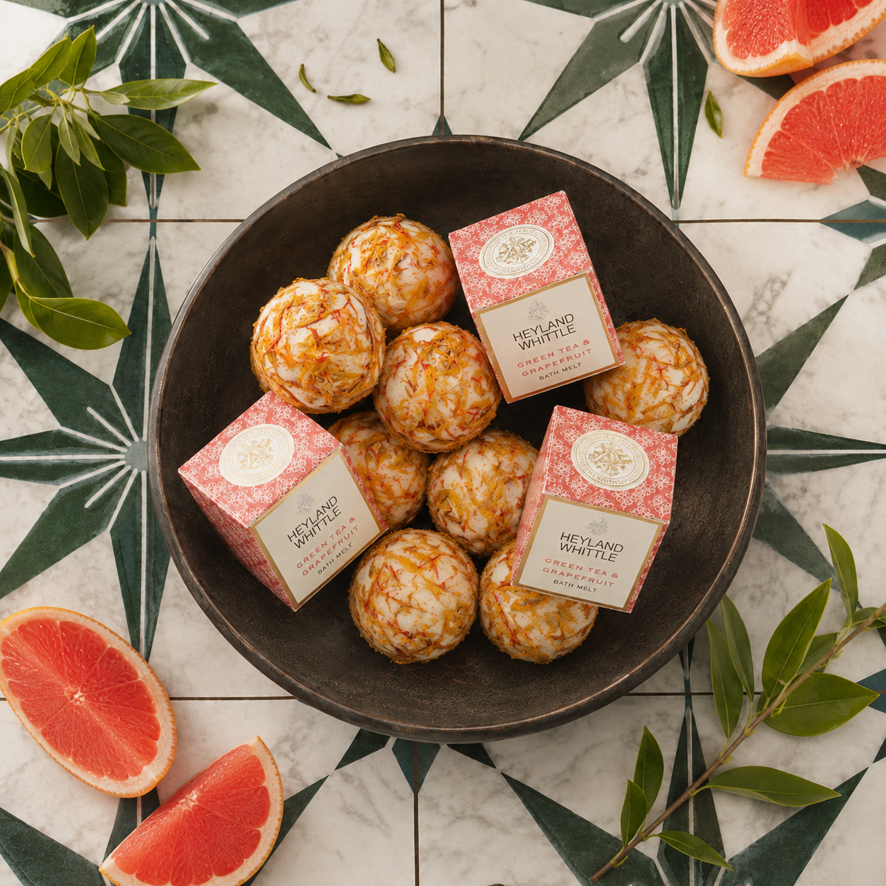Two pink and cream packaged bath bombs labeled 'HEYLAND & WHITTLE' arranged in a dark bowl with several wrapped bath bombs, styled on a white marble surface with green star-pattern tiles, grapefruit slices, and green leaves.