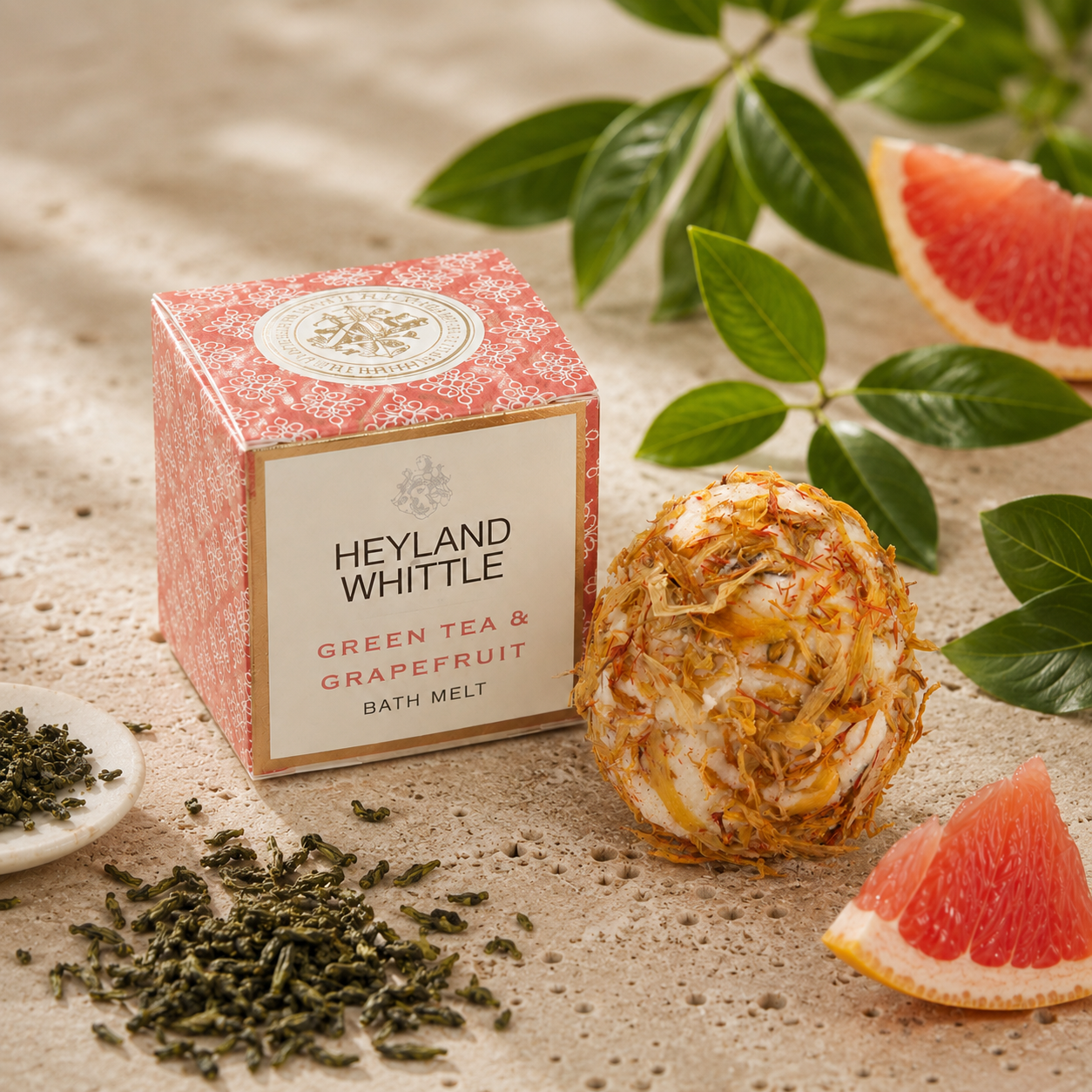 Heyland Whittle Green Tea & Grapefruit Bath Melt box shown beside a wrapped bath melt ball, with loose tea leaves, grapefruit slices, and green leaves on a textured beige surface.