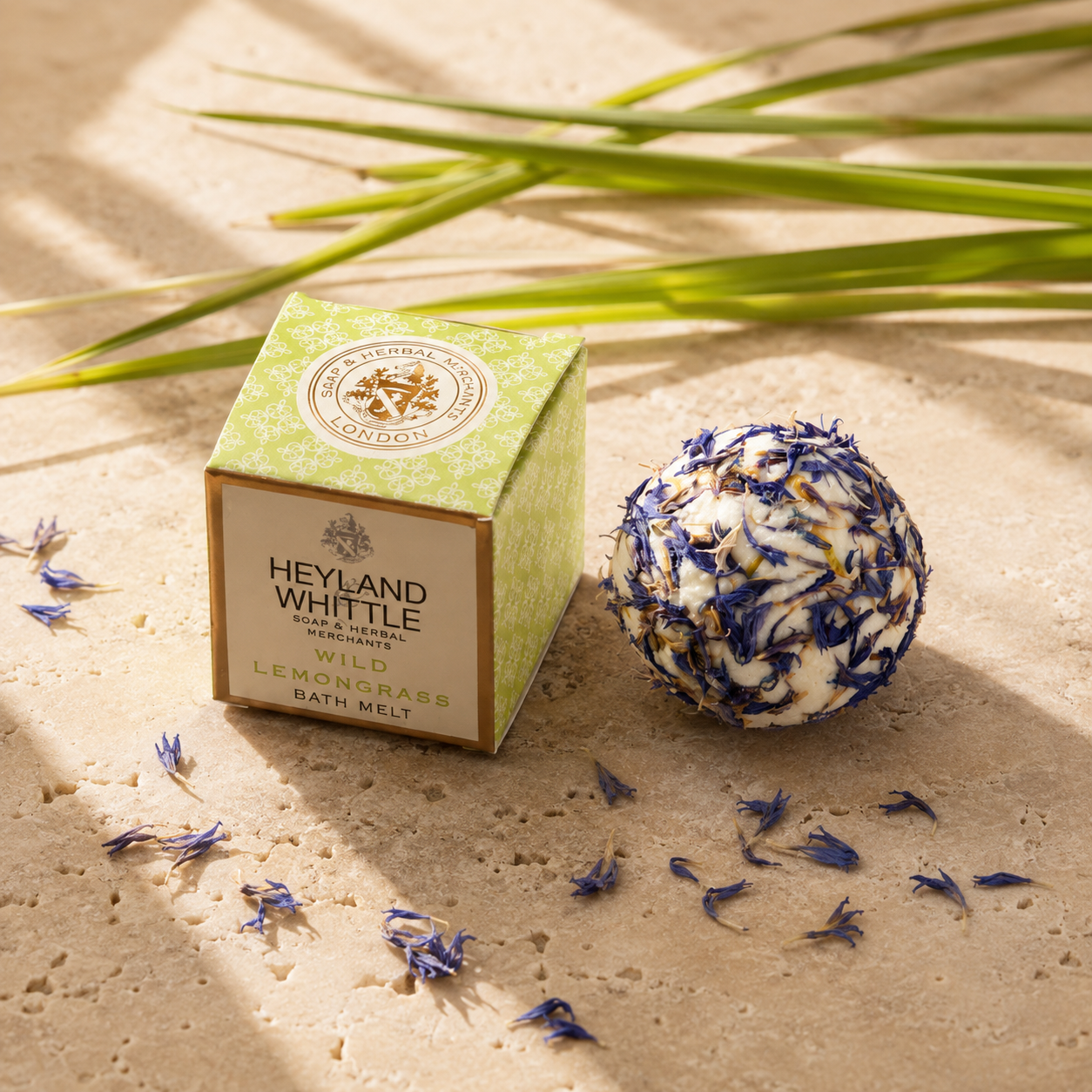 Heyland Whittle Wild Lemongrass Bath Melt shown with its green and beige branded box and a round bath bomb made of dried botanicals, styled on a sandy surface with scattered flower petals and green leaves.