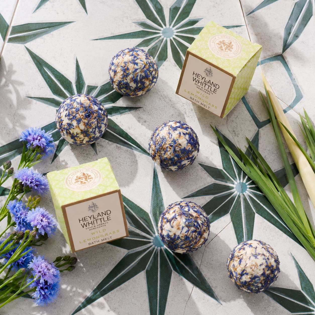 Five round lavender and cream bath salts with blue dried flower inclusions are arranged on a patterned surface with two Heyland Whittle boxes, blue flowers, and green leaves.