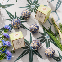 Five round lavender and cream bath salts with blue dried flower inclusions are arranged on a patterned surface with two Heyland Whittle boxes, blue flowers, and green leaves.