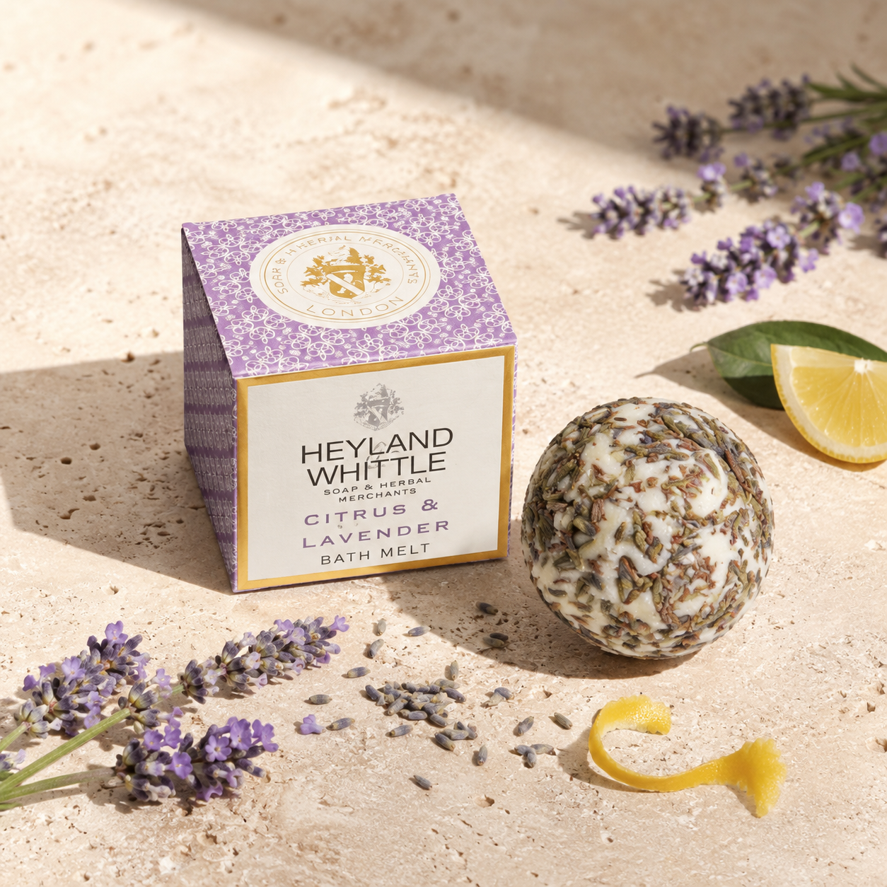 A boxed bath melt and a round bath melt ball are styled on sandy ground with lavender flowers, a lemon slice, and a lemon peel. The box reads Heyland Whittle Citrus & Lavender Bath Melt.