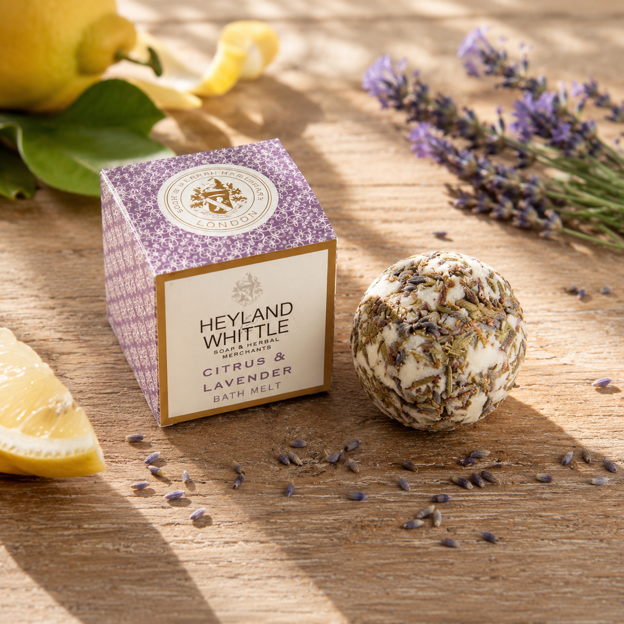 Heyland Whittle London Citrus & Lavender Bath Melts shown as a purple patterned box next to a dried lavender and citrus bath bomb, styled on a wooden table with lemon slices and lavender sprigs.