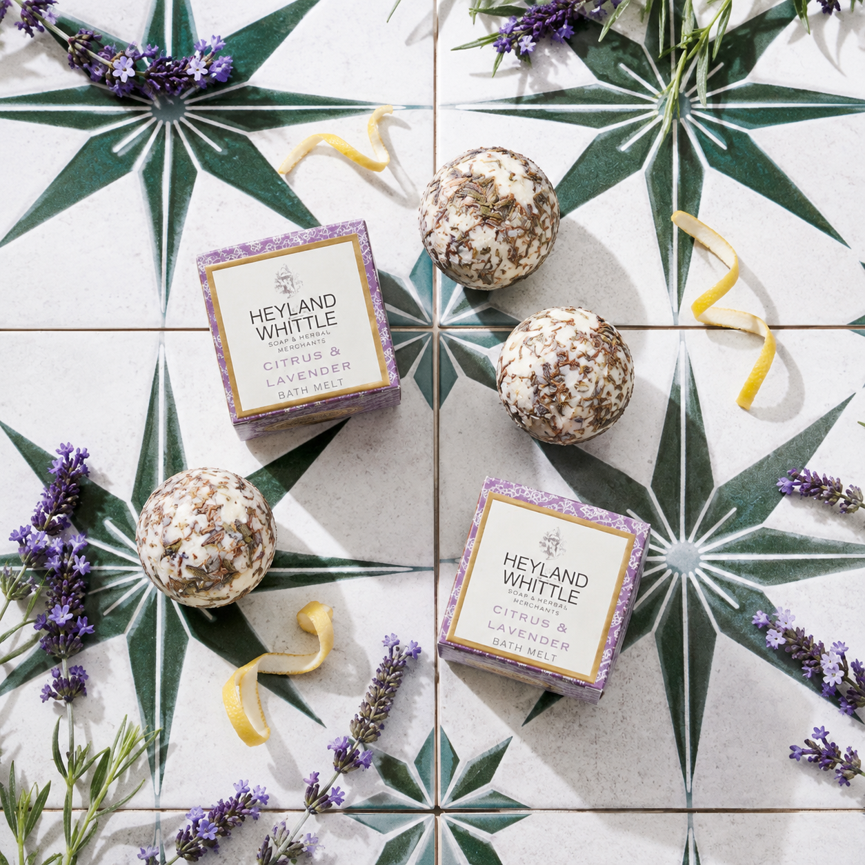 Three Heyland Whittle citrus and lavender hand wash mists are arranged on a white tiled surface with green star-patterned tiles, surrounded by lavender sprigs, lemon curls, and dried botanicals. Two matching purple and cream boxes are also visible.