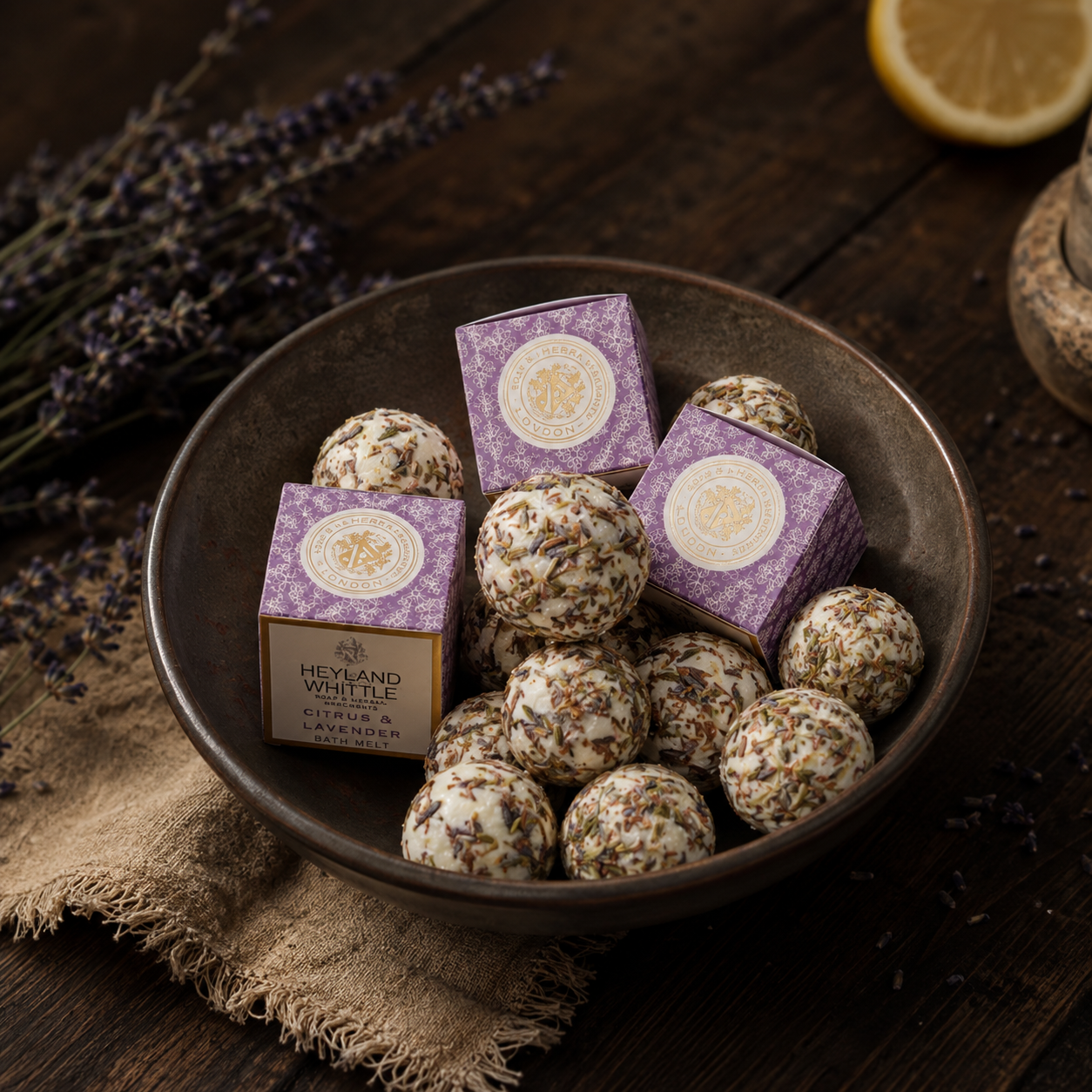 A bowl filled with lavender-infused white chocolate truffles and two purple branded boxes, styled on a dark wooden surface with dried lavender, burlap, and a lemon slice.