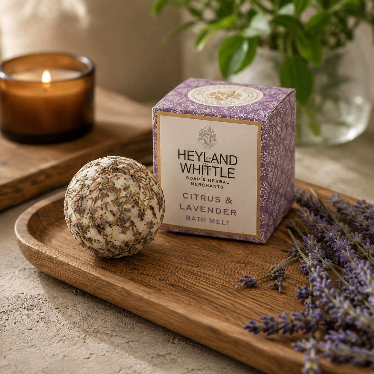 A bath product scene showing a round lavender-infused bath bomb next to its purple patterned box labeled Heyland Whistle Citrus & Lavender Bath Melt, styled on a wooden tray with dried lavender and a lit candle in the background.