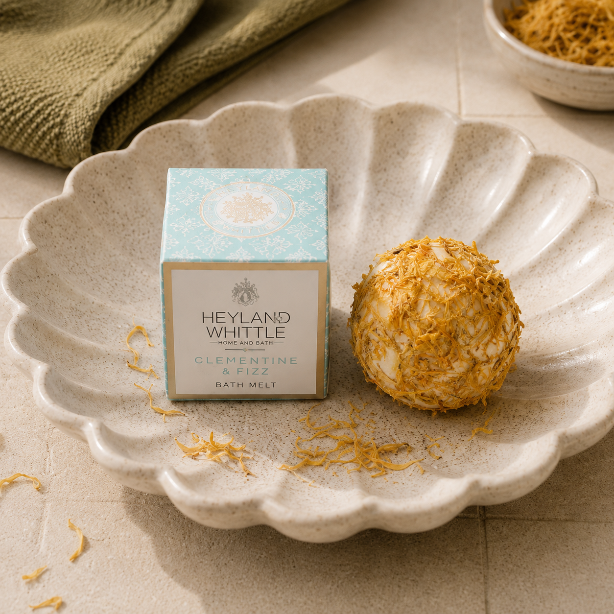A boxed bath melt product sits on a scalloped ceramic plate beside a round bath bomb coated in dried citrus petals. The box reads Heyland  Whittle and Clementine & Fizz Bath Melt.