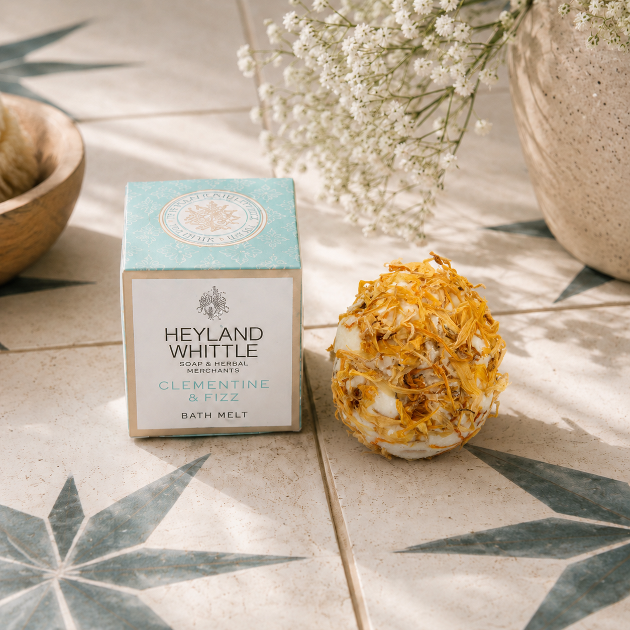 Heyland Whittle Clementine & Fizz bath melt shown with its light blue and white box next to a dried citrus peel sphere, styled on a tiled surface with baby's breath flowers.