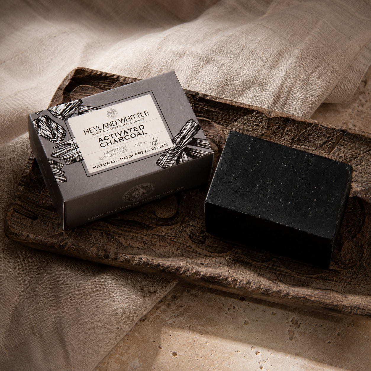 A dark charcoal soap bar beside its gray box packaging, styled on a rustic wooden tray with draped linen fabric in warm natural light. The box reads 'HERB AND WHITTLE ACTIVATED CHARCOAL SOAP'.
