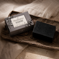 A dark charcoal soap bar beside its gray box packaging, styled on a rustic wooden tray with draped linen fabric in warm natural light. The box reads 'HERB AND WHITTLE ACTIVATED CHARCOAL SOAP'.