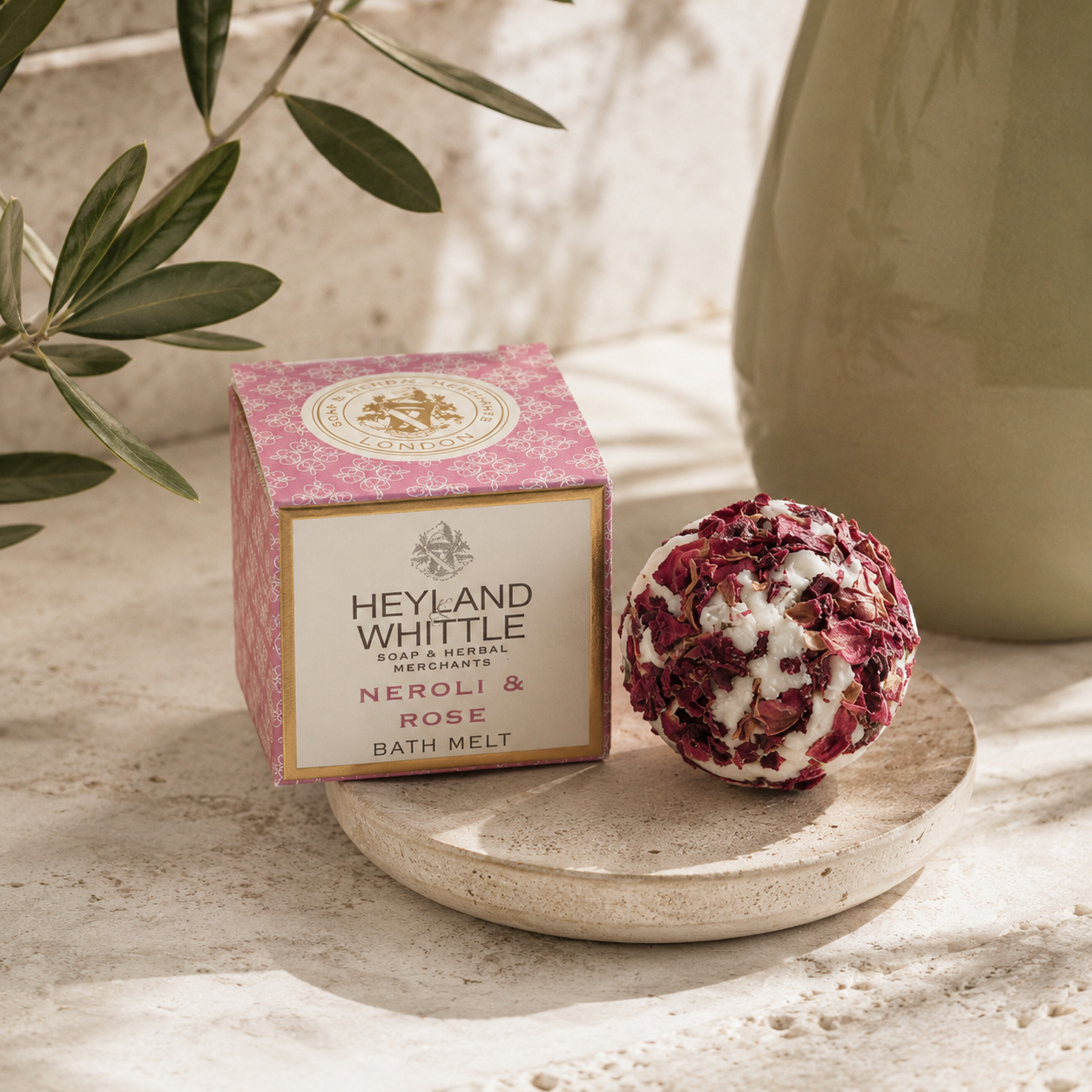 Heyland Whittle Neroli & Rose Bath Melt shown with its pink patterned box and a round bath bomb made of dried rose petals and white wax, styled on a stone surface with olive branches and a ceramic vase.