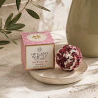 Heyland Whittle Neroli & Rose Bath Melt shown with its pink patterned box and a round bath bomb made of dried rose petals and white wax, styled on a stone surface with olive branches and a ceramic vase.