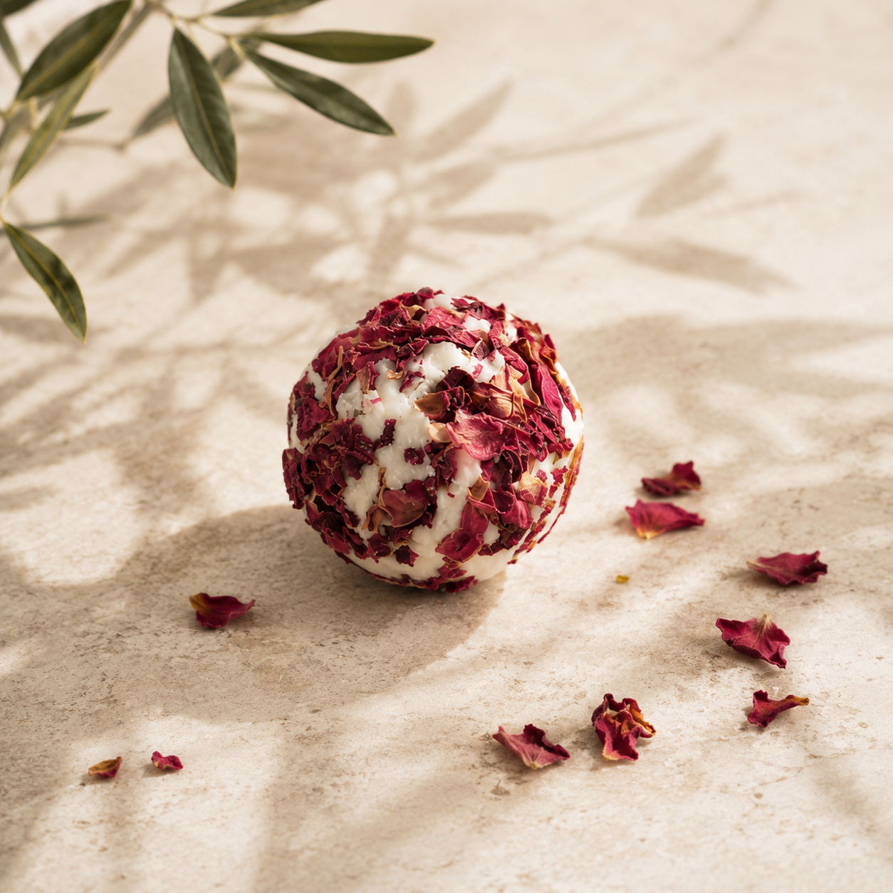 A spherical bath bomb made of dried red rose petals and white wax, placed on a textured beige surface with olive leaves and scattered rose petals under soft shadows.
