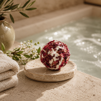 A spherical bath bomb made of dried red roses and white wax sits on a round stone dish beside a folded towel, with a spa pool in the background.