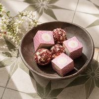Four pink patterned gift boxes and four round pink-and-white decorative truffles arranged in a dark ceramic bowl on a geometric tile surface with white flowers nearby.
