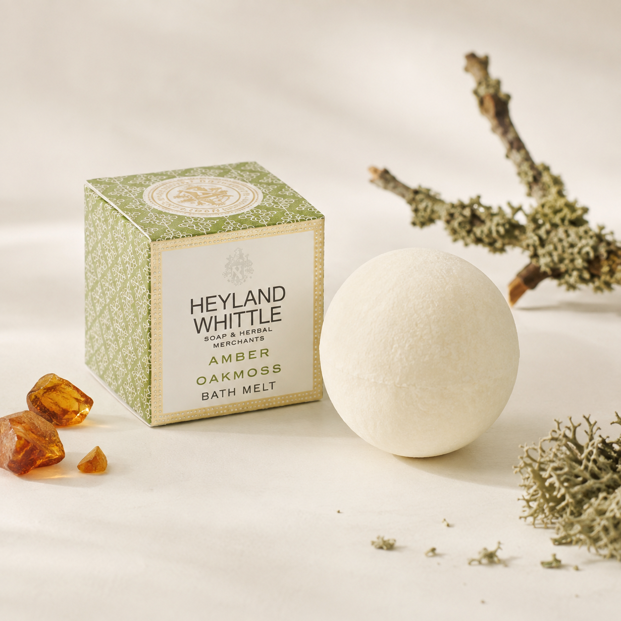 A bath melt product shown with its branded box and a round off-white bath bomb on a soft cream surface, styled with amber crystals, dried branches, and botanicals. The box reads Heyland Whittle, Soap & Herbal Merchants, Amber Oakmoss Bath Melt.