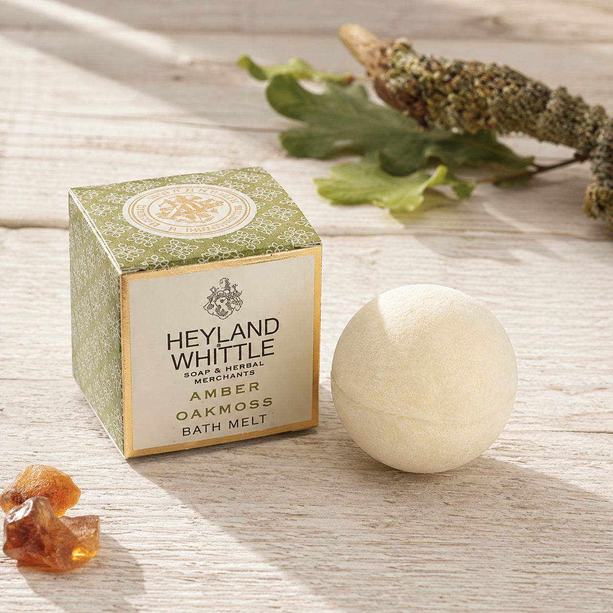 Heyland Whittle Amber Oakmoss bath melt shown with its textured green and gold box next to a round off-white bath bomb, styled with amber crystals and dried botanicals on a light wood background.