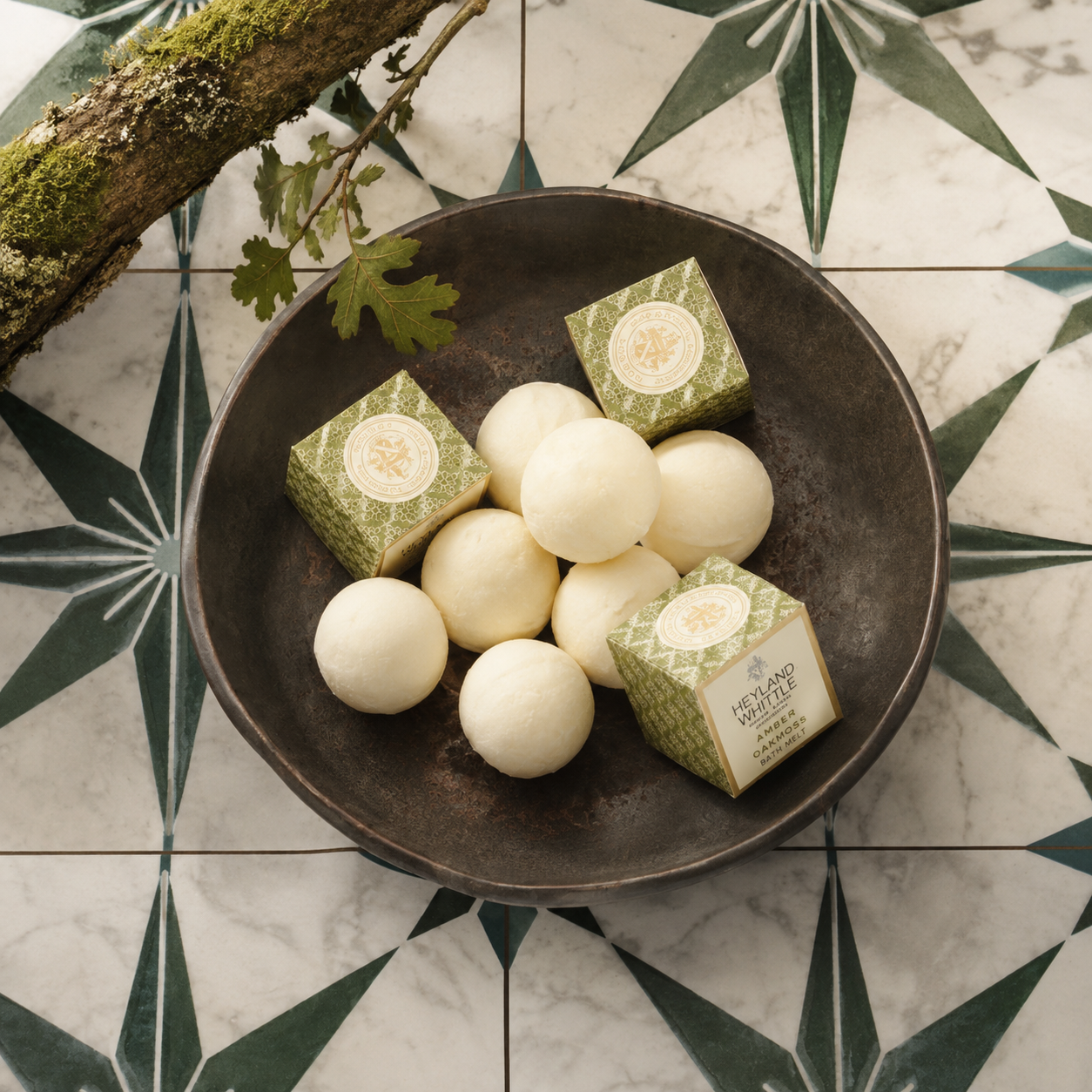 A top-down view of white soap bars arranged in a dark metal bowl, with two green patterned boxes of Heyland & Whittle. on a decorative black and white tiled surface.