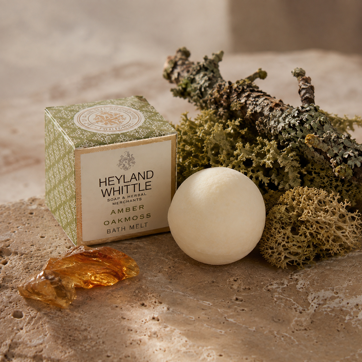 A round off-white bath melt bar sits beside its patterned green and cream box labeled Heyland Whittle Amber Oakmoss Bath Melt, styled with dried moss, bark, and amber pieces on a stone surface.