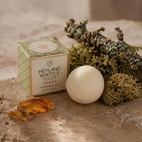 A round off-white bath melt bar sits beside its patterned green and cream box labeled Heyland Whittle Amber Oakmoss Bath Melt, styled with dried moss, bark, and amber pieces on a stone surface.