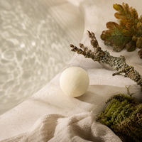 A round, off-white bath bomb resting on a textured beige fabric surface next to a branch, moss, and a leaf, with a blurred background of rippling water.