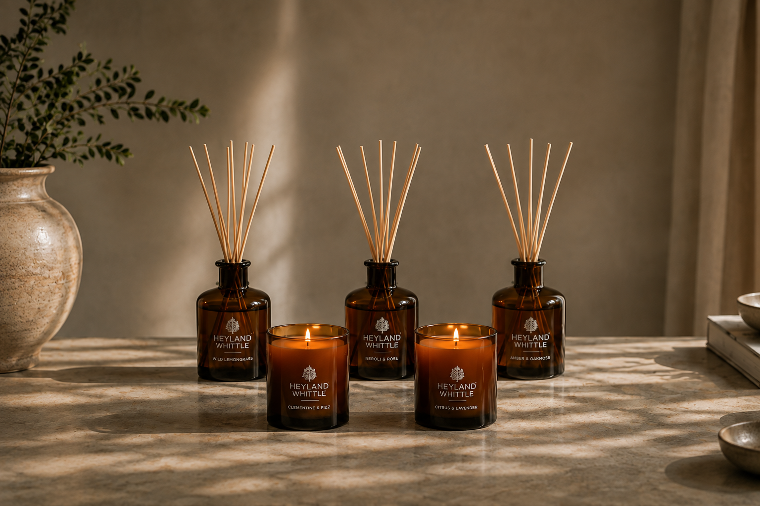 Four Heyland & Whittle reed diffusers and two matching amber glass candles arranged on a tabletop in warm natural light, with a vase and books softly blurred in the background.
