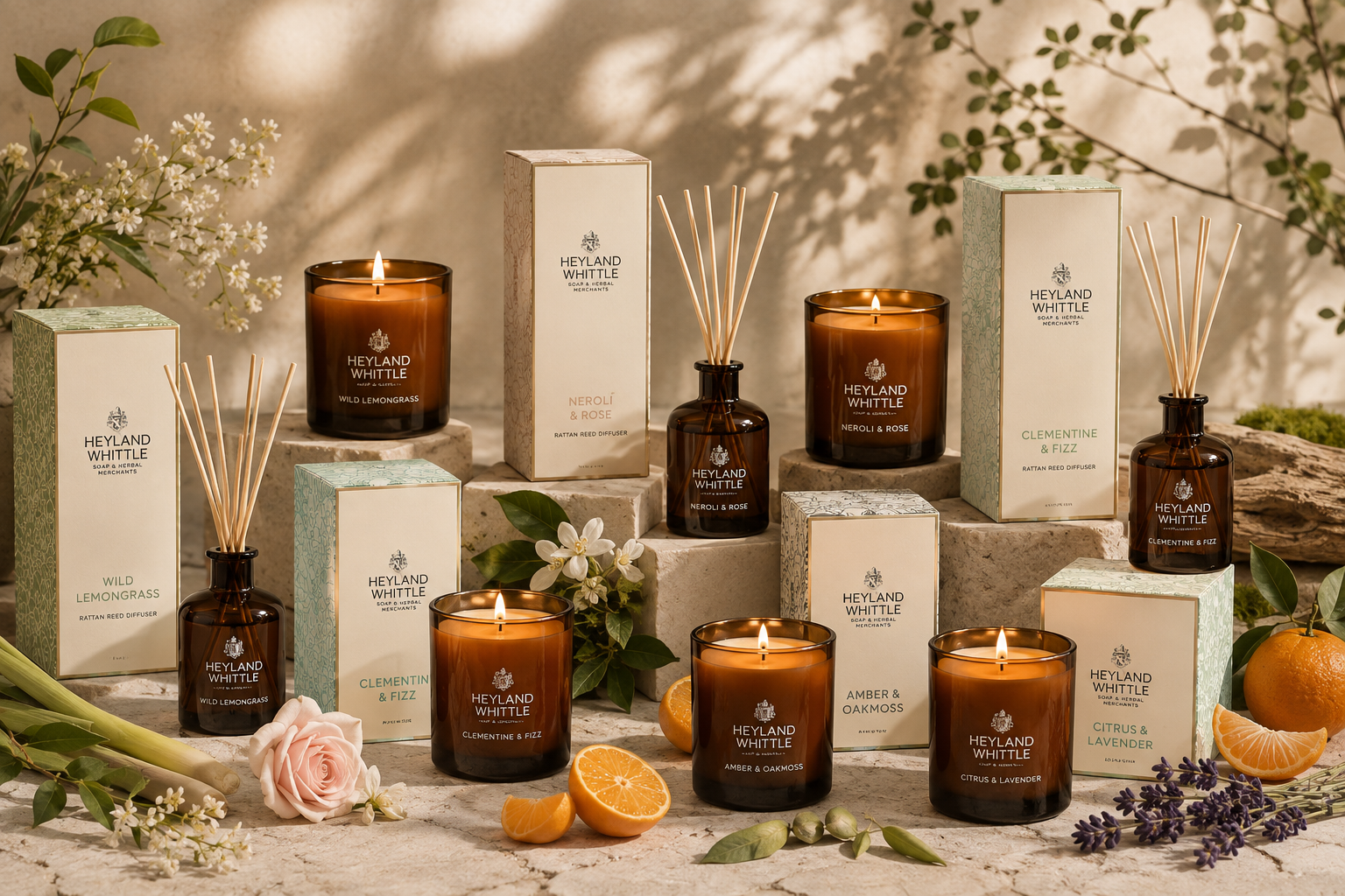 A styled product display of Heyland Whittle home fragrance products, including amber glass candles, reed diffusers, and matching boxes, arranged on stone blocks with flowers, citrus, and greenery in a warm, natural setting.