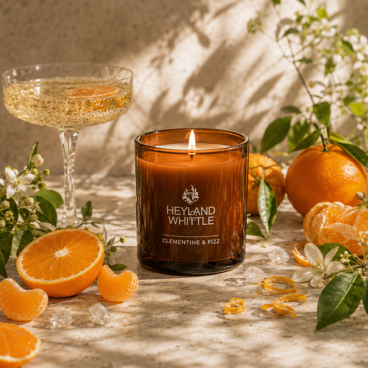 An amber glass scented candle labeled 'HEYLAND WHITTLE CLEMENTINE & FIZZ' sits lit on a sunlit stone surface, surrounded by oranges, orange slices, white flowers, ice, and a coupe glass of sparkling drink.