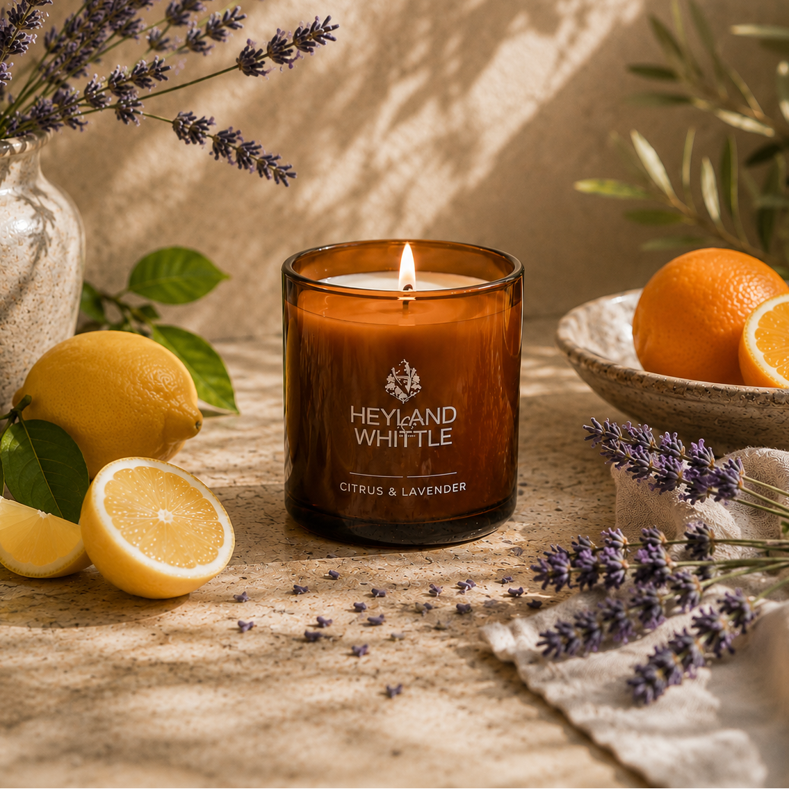 A lit amber glass candle jar labeled Heyland Whittle Citrus & Lavender, styled on a linen surface with lemons, oranges, lavender sprigs, and a ceramic bowl in a warm rustic setting.