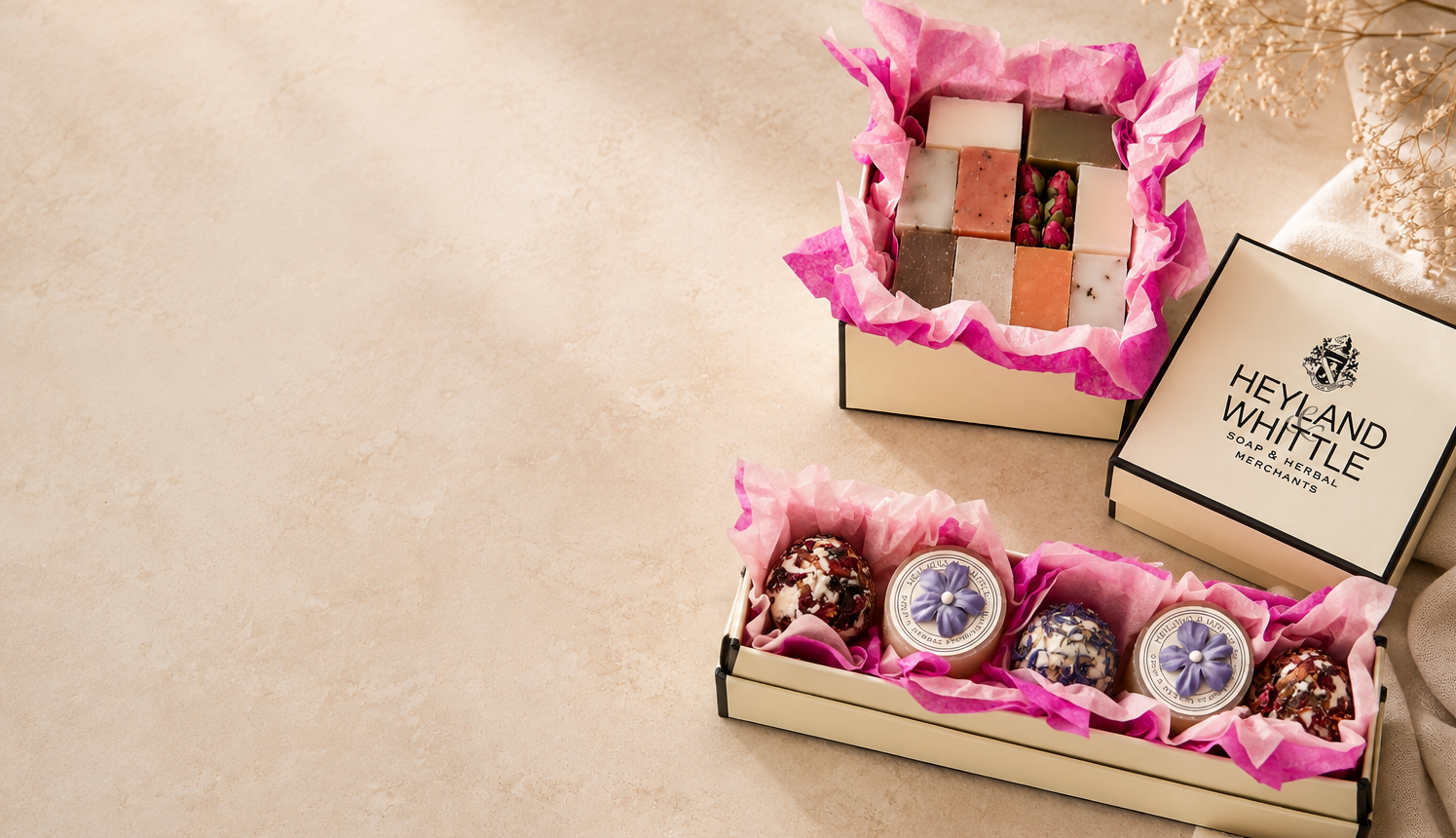 A gift box of assorted chocolates and sweets from Heyland Whittle, shown open with pink tissue paper on a beige surface. One box contains a grid of assorted chocolates, and another open box at the bottom shows three decorated round chocolates.