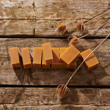 Several rectangular bars of warm amber-orange soap arranged on a rustic wooden surface, with dried thistle stems placed around them.