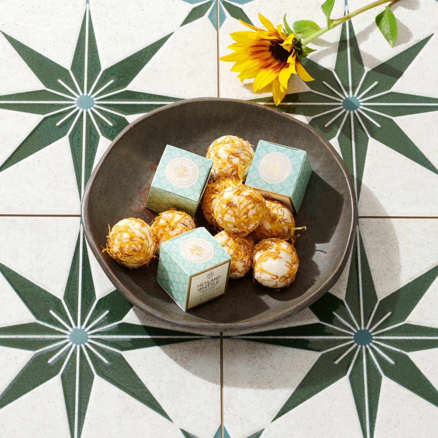 A dark bowl filled with bath melts and two small branded boxes sits on a white tiled surface with a green starburst pattern and a yellow sunflower nearby.