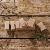 Several rectangular bars of natural beige soap arranged on a rustic wooden surface, with dried botanical sprigs placed around them.