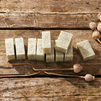 Several rectangular bars of speckled soap arranged on a rustic wooden surface, with dried flower pods placed nearby as styling props.