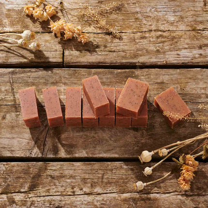 Several rectangular bars of dusty rose pink soap with dark flecks are arranged on a rustic wooden surface, surrounded by dried flowers and botanicals.