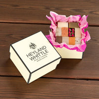 A boxed assortment of handmade soaps displayed on a dark wood surface. One cream-colored box lid is shown beside an open box lined with bright pink tissue paper, revealing multiple square soaps in natural tones and a small pink-and-red patterned bar.