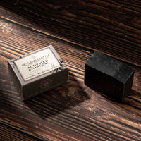 A bar of black activated charcoal soap sits next to its dark gray packaging box on a dark rustic wooden surface. The box is labeled 'HEYLAND WHITTLITE ACTIVATED CHARCOAL SOAP'.