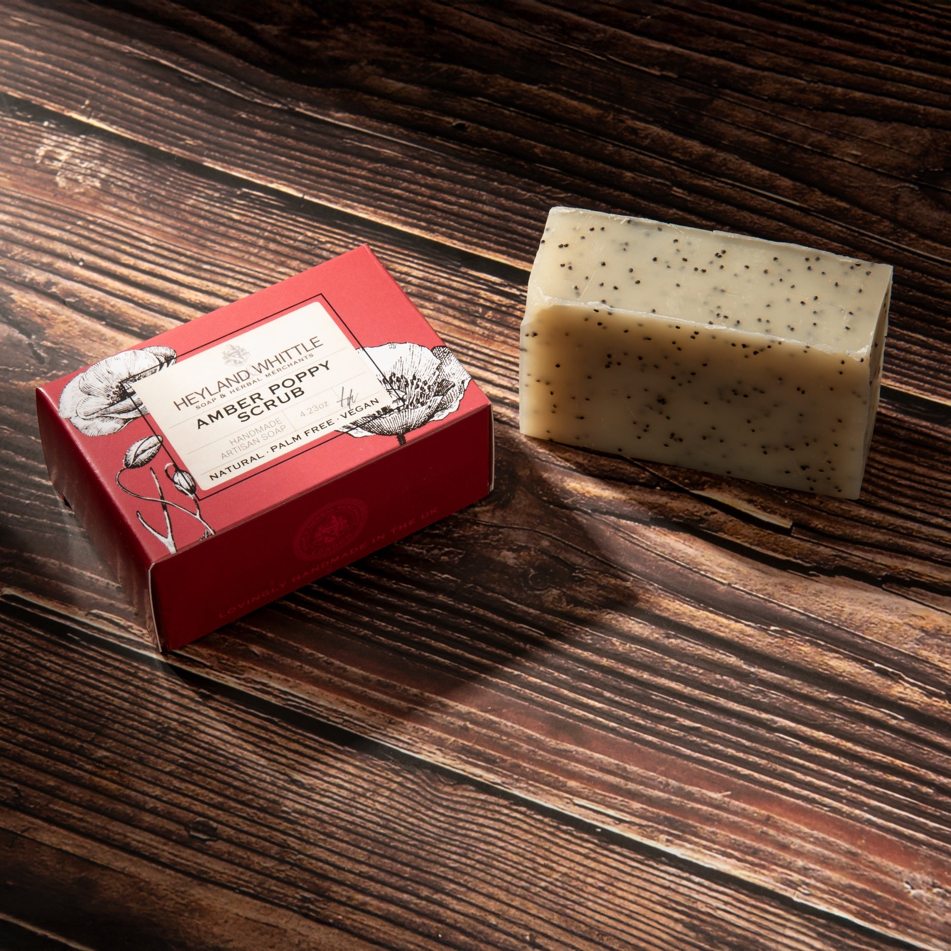 A rectangular bar of Heyland & Whittle 'Amber Poppy Scrub' soap sits on a dark wooden surface next to its red packaging box.