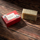 A rectangular bar of Heyland & Whittle 'Amber Poppy Scrub' soap sits on a dark wooden surface next to its red packaging box.