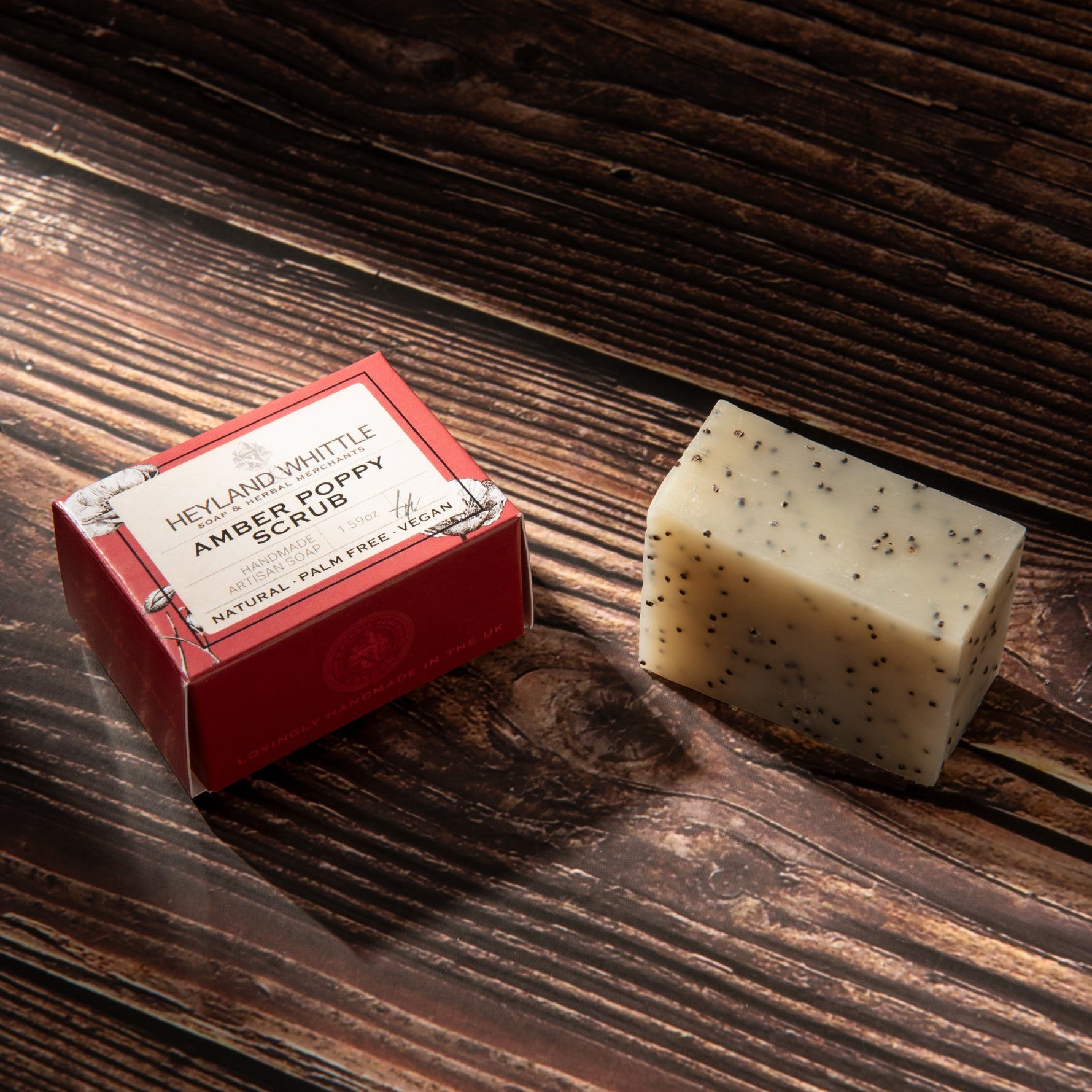 A bar of Heyland & Whittle Amber Poppy Scrub soap next to its red packaging box, displayed on a dark rustic wooden surface.